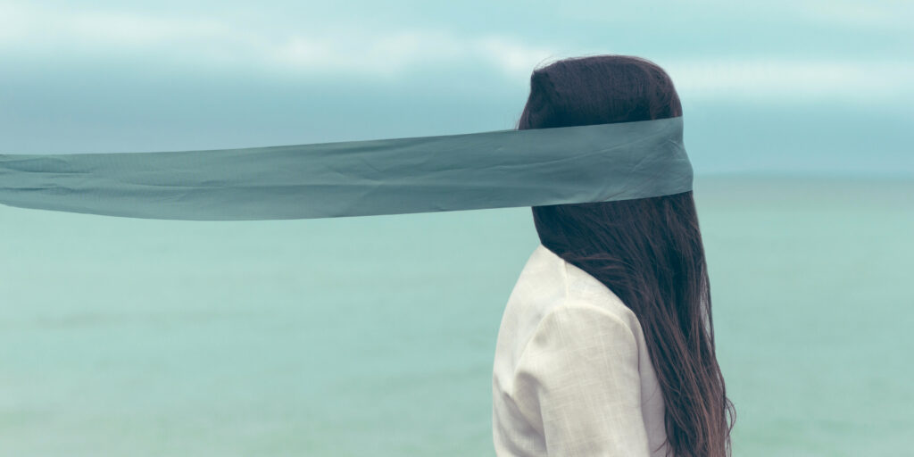 Woman by the sea blindfolded with a long outstretched piece of material.