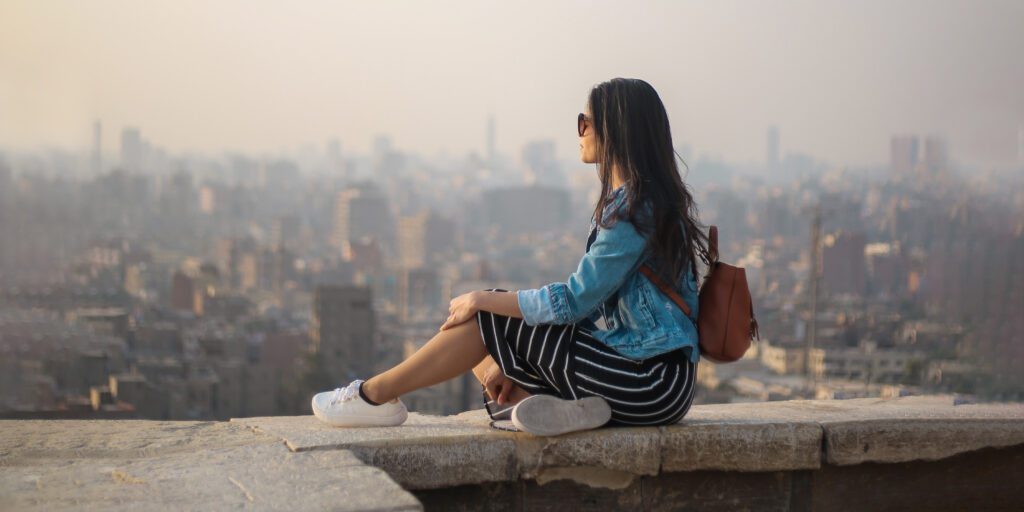 Woman with a backpack and sunglasses sitting on a wall overlooking the city from above.