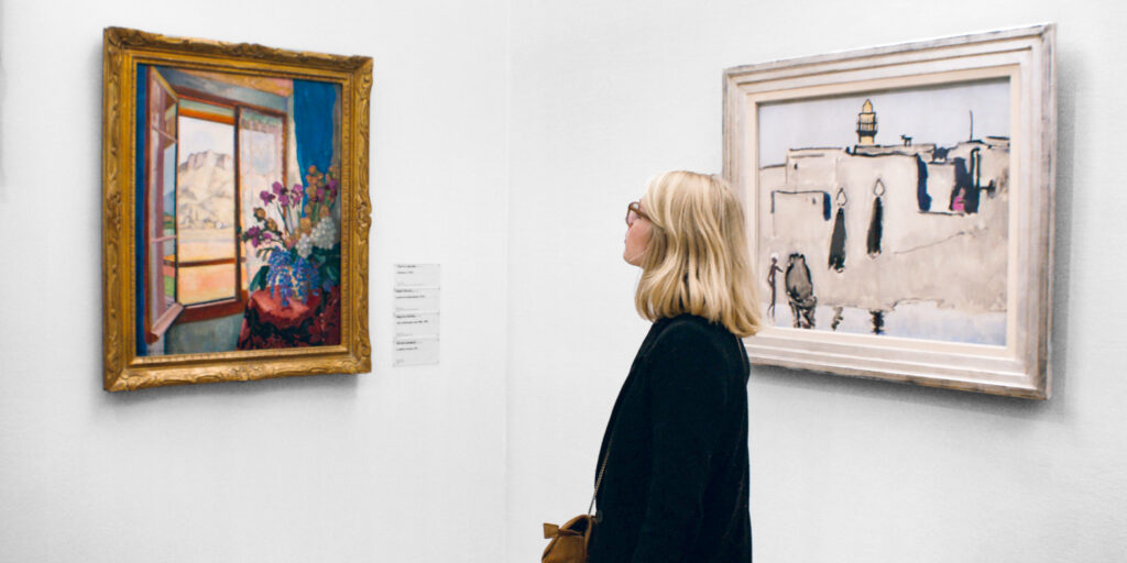 Blonde woman with glasses looking at paitnings hanging on the wall in a gallery.