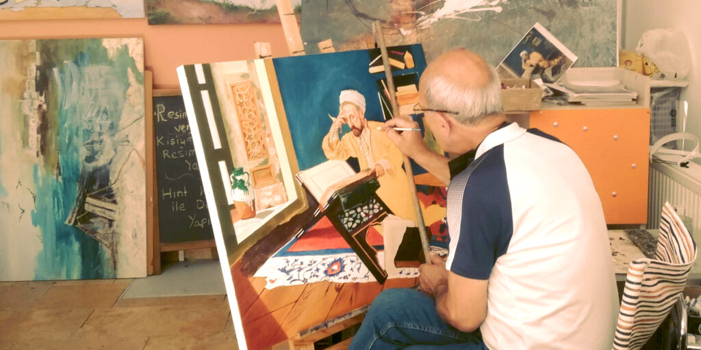Old man sitting down painting in his studio.