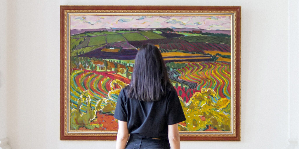 A woman standing in front of a large, hanging, and very colourful landscape painting.