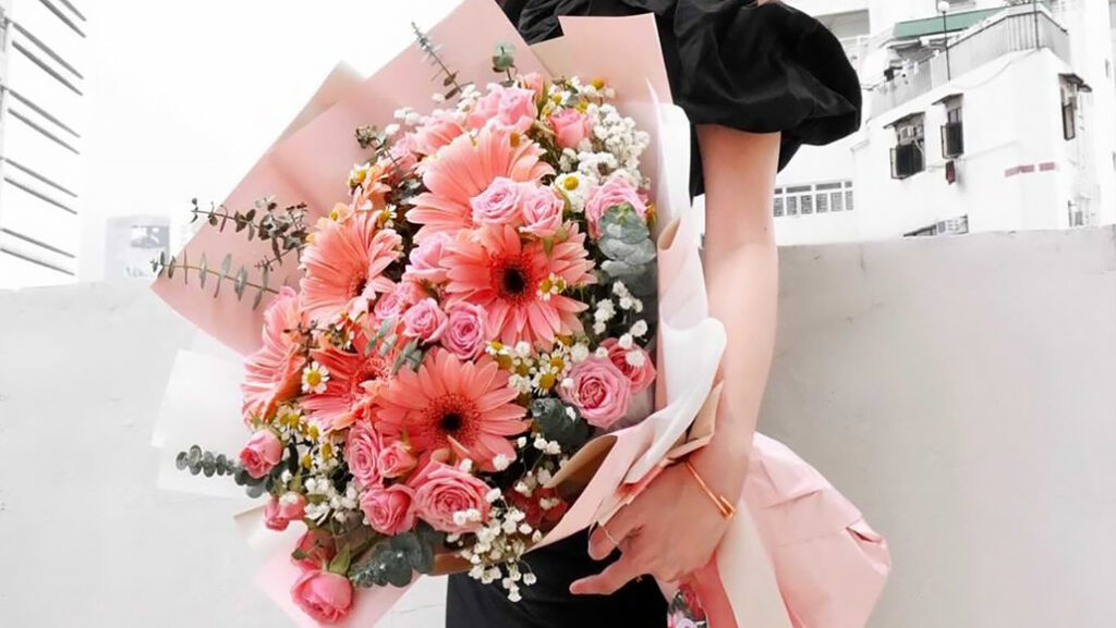 Woman in a black dress holding a big bouquet of different pink and peach coloured flowers wrapped in pink tissue paper and tied with a ribbon.