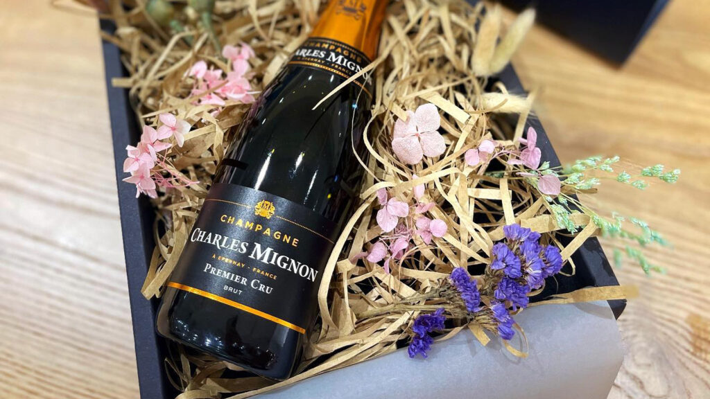 A bottle of champagne in an open gift box with straw and dried flowers.